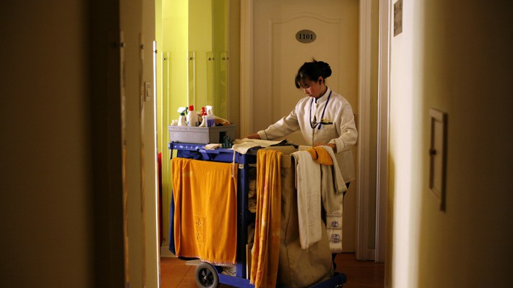 A hotel maid enters a room with a cart of cleaning supplies.