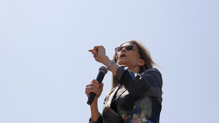 Marianne Williamson speaks into a microphone at the Iowa State Fair. She's wearing sunglasses and a watch, and the blue sky is visible behind her.