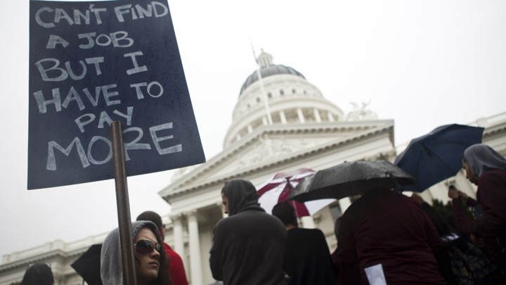 Woman stands with a sign that reads "Can't find a job but I have to pay more" in California