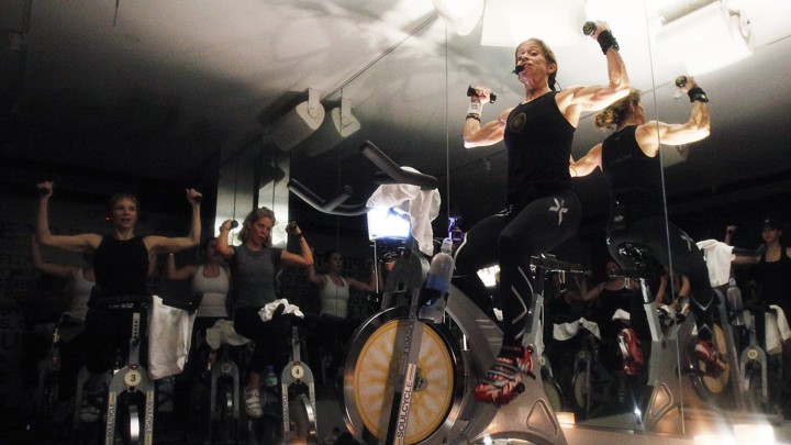 A woman lifts weights while riding a SoulCycle stationary bicycle.