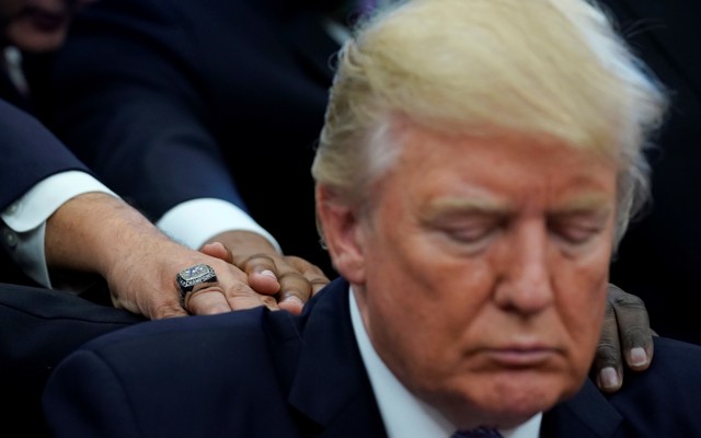 Donald Trump looks down as three men, whose faces are not visible, lay their hands on his back and shoulders.