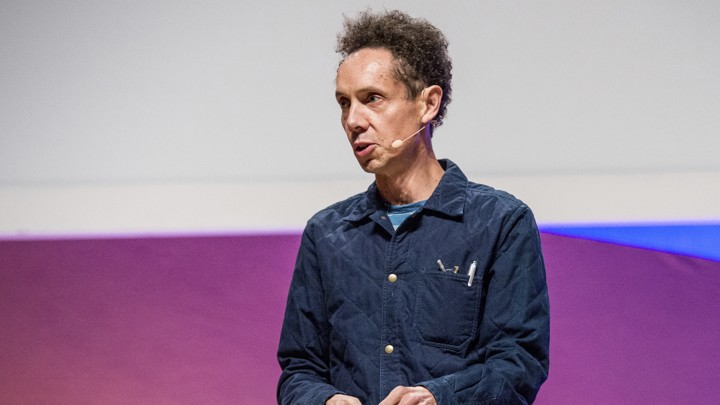Malcolm Gladwell stands in front of a purple-and-blue screen giving a speech.
