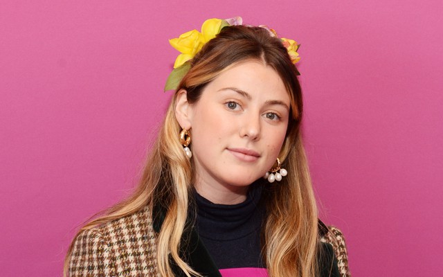 Caroline Calloway stands in front of a pink backdrop looking straight into the camera.