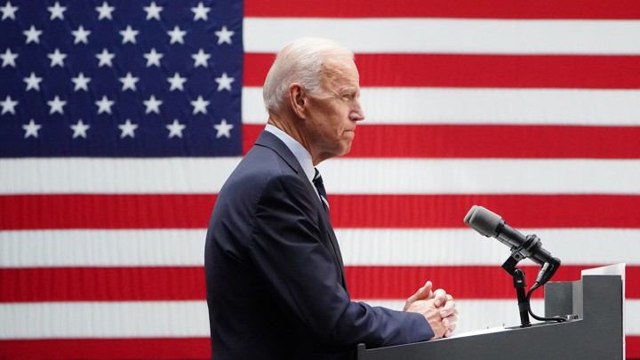 Standing in front of a podium, Joe Biden pauses while delivering remarks into a microphone. A large American flag is visible behind him.