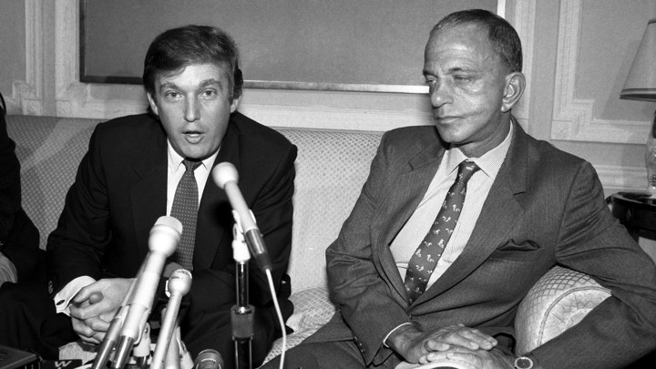 A black and white photo of Donald Trump sitting in front of microphones next to Roy Cohn.