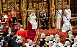 Queen Elizabeth and Prince Charles sit on golden thrones, surrounded by members of Parliament and other officials in both formal and official attire..