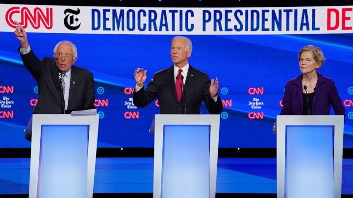 Vermont Senator Bernie Sanders, standing next to former Vice President Joe Biden and Senator Elizabeth Warren of Massachusetts, all behind lecterns on a stage, raises his hand during the fourth Democratic primary debate.