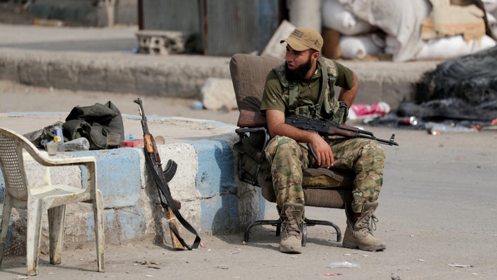 A man dressed in army fatigues sits on a chair holding a gun. Trash is strewn behind him, and an empty white chair and a gun are next to him.