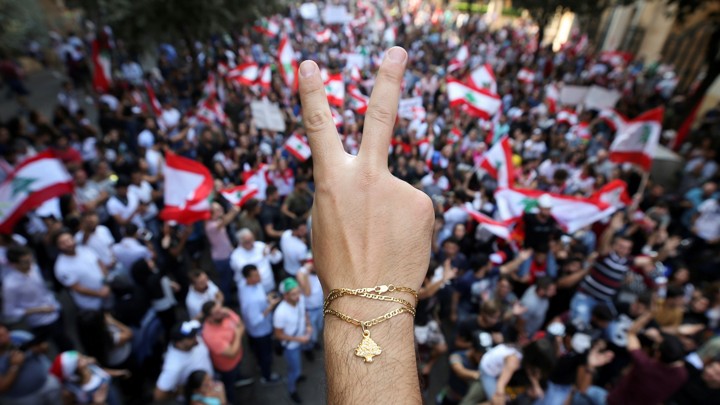 A close up photo of a demonstrator flashing a V sign. In the background, protesters gather waving Lebanon flags.