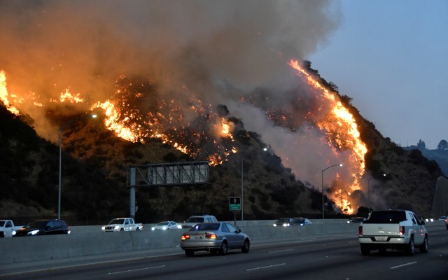 A fire burns on a hillside next to a freeway as cars drive by.