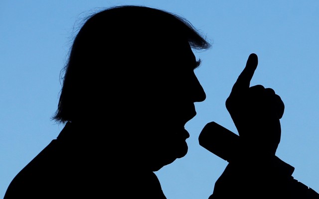 A silhouette of Donald Trump talking into a microphone with his finger pointing to the sky.