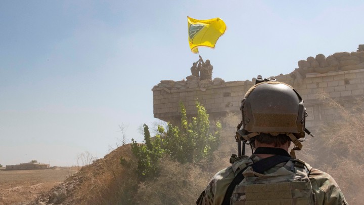 A U.S. soldier watches Syrian Democratic Forces raise a flag in the background.