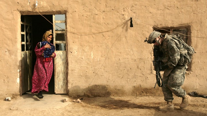 An Iraqi woman, carrying a newborn infant, leaves her home as a U.S. Army soldier prepares to search the house.
