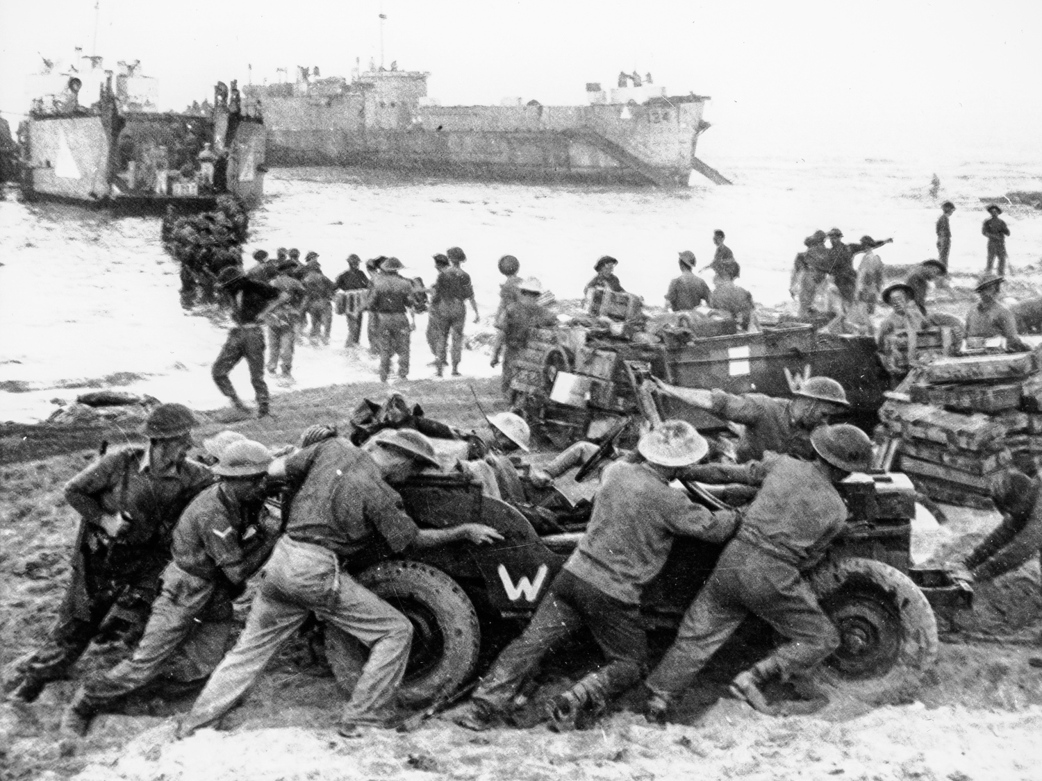 Allied soldiers push a jeep while others unload military equipment and ...