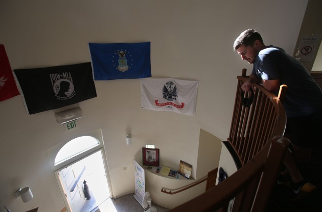 A man stands at the top of staircase overlooking a lobby. There are POW-MIA flags on the wall to his right.