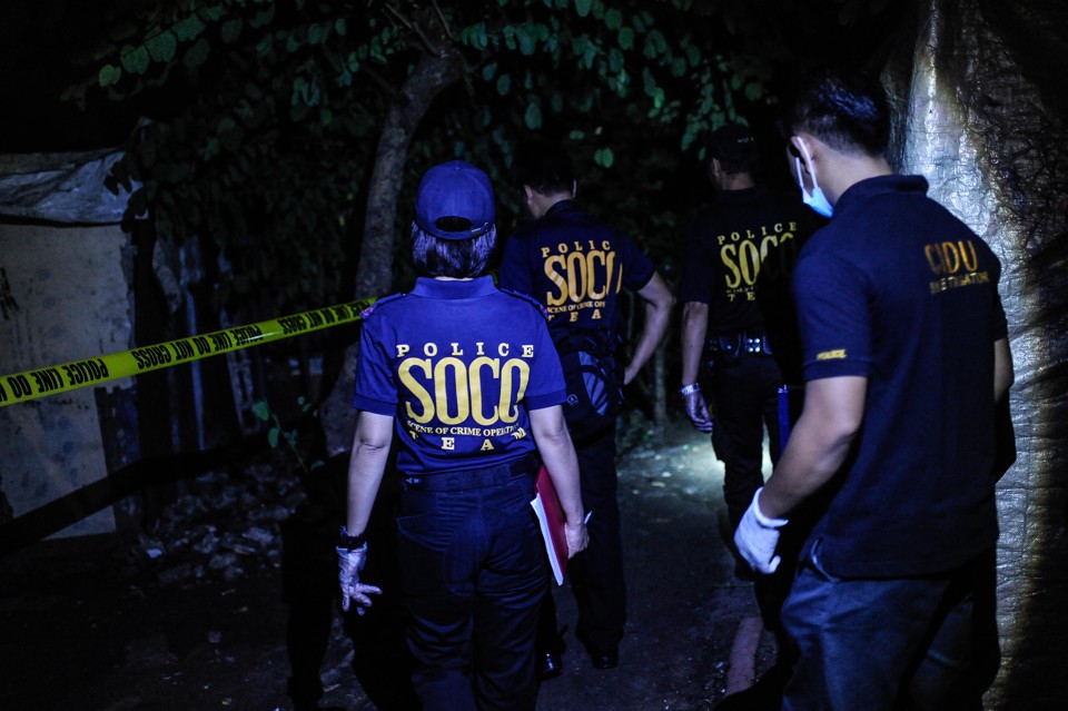 Police officers inspect evidence amongst trees and police tape.