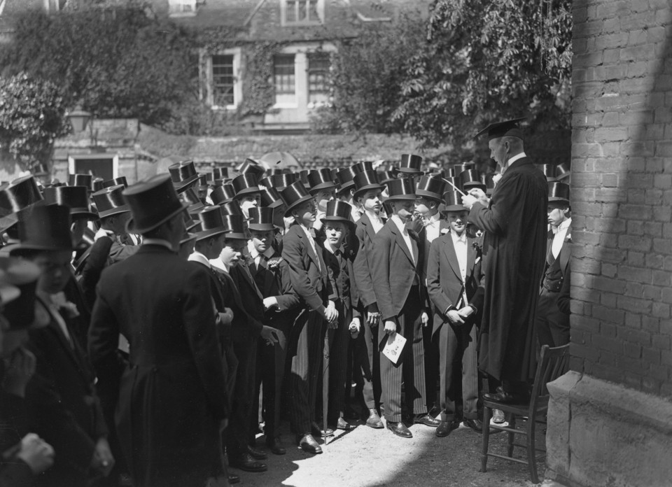 Etonians wearing hats and tails gather as a schoolmaster reads from the registrar.