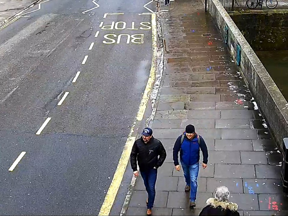 Two men wearing hats and jackets walk down a sidewalk, as captured on CCTV footage.