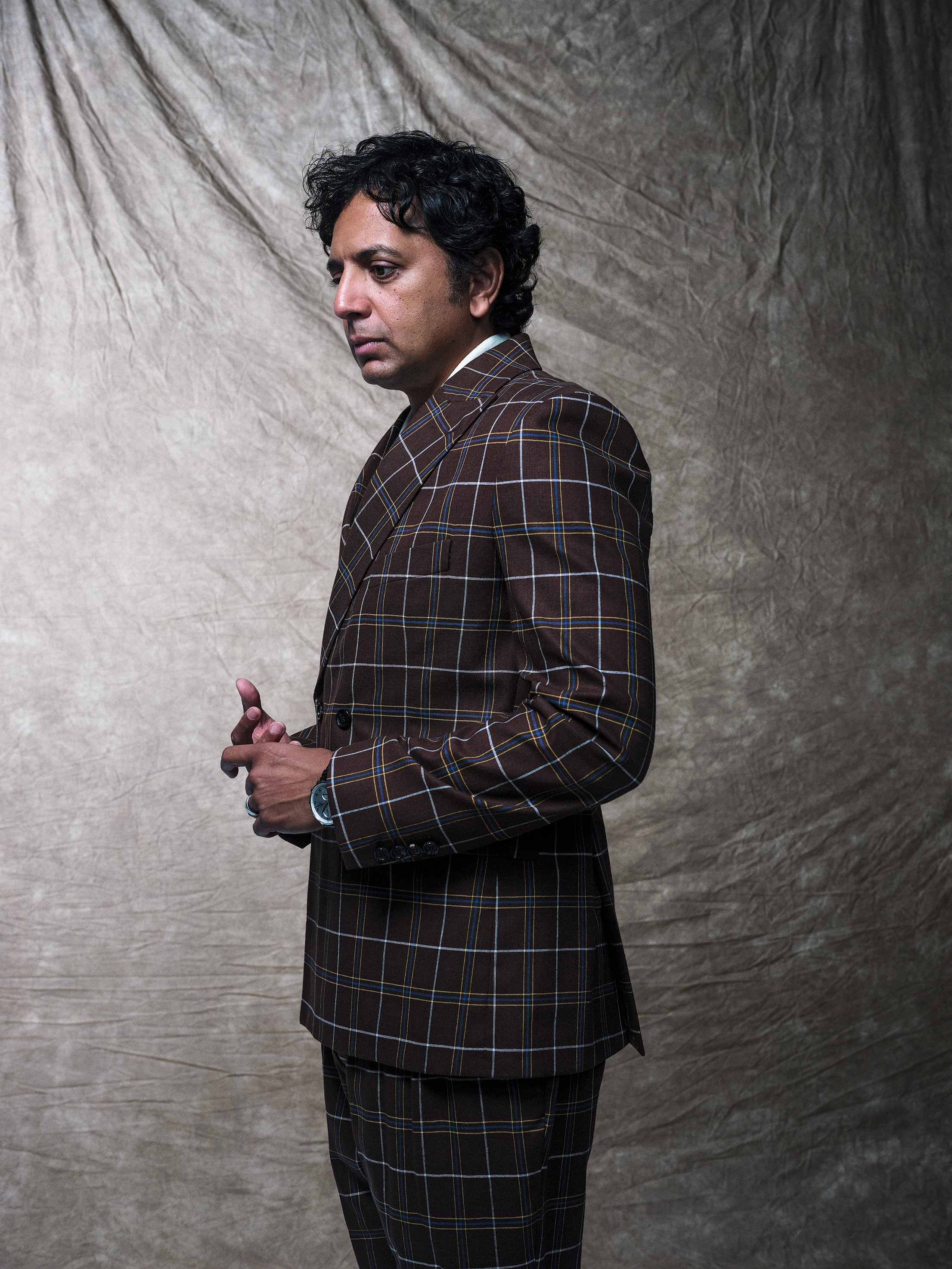 portrait of M. Night Shyamalan standing, looking down with hands clasped, wearing brown plaid suit