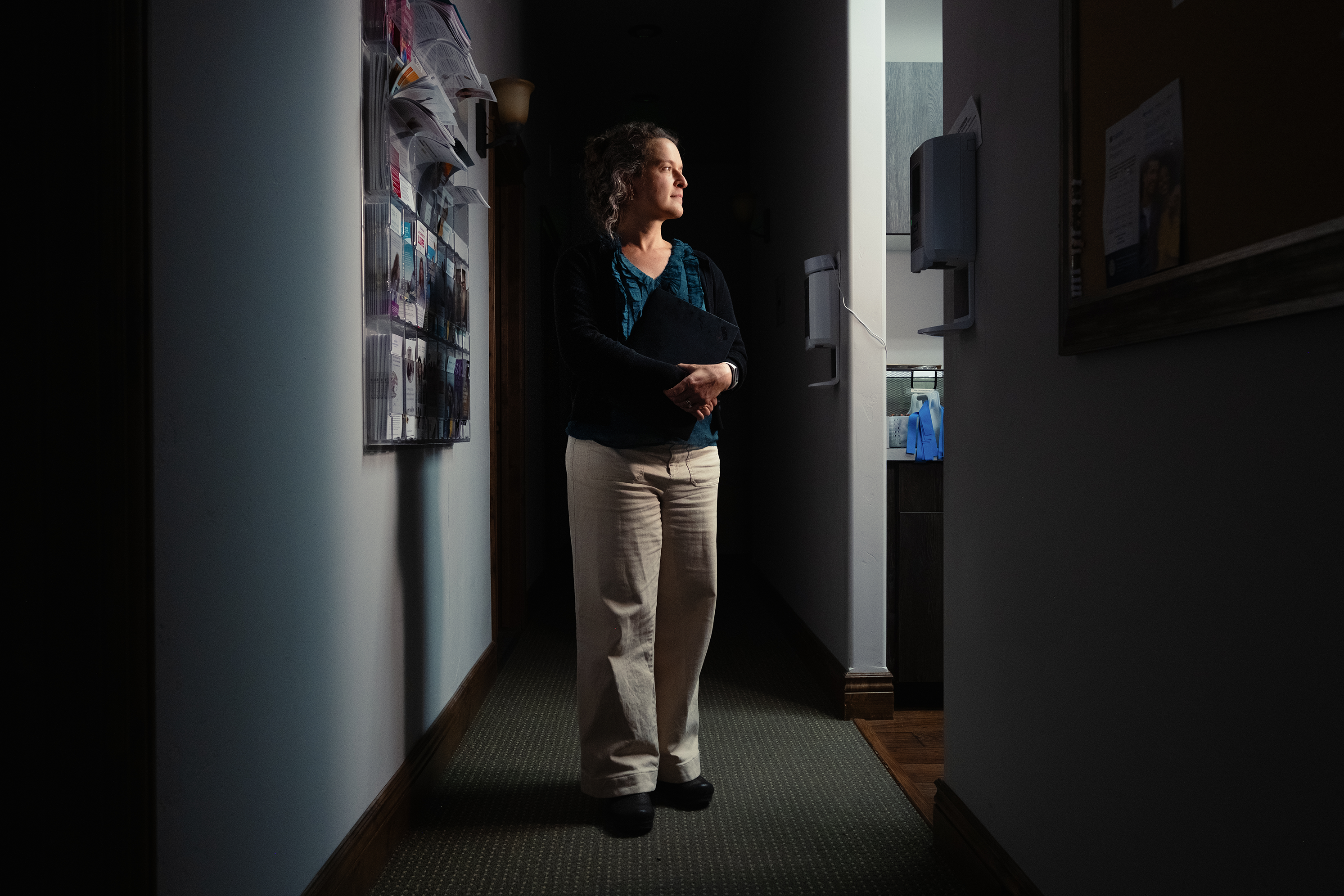 A photo of woman standing in shadowy hallway next to a patient exam room