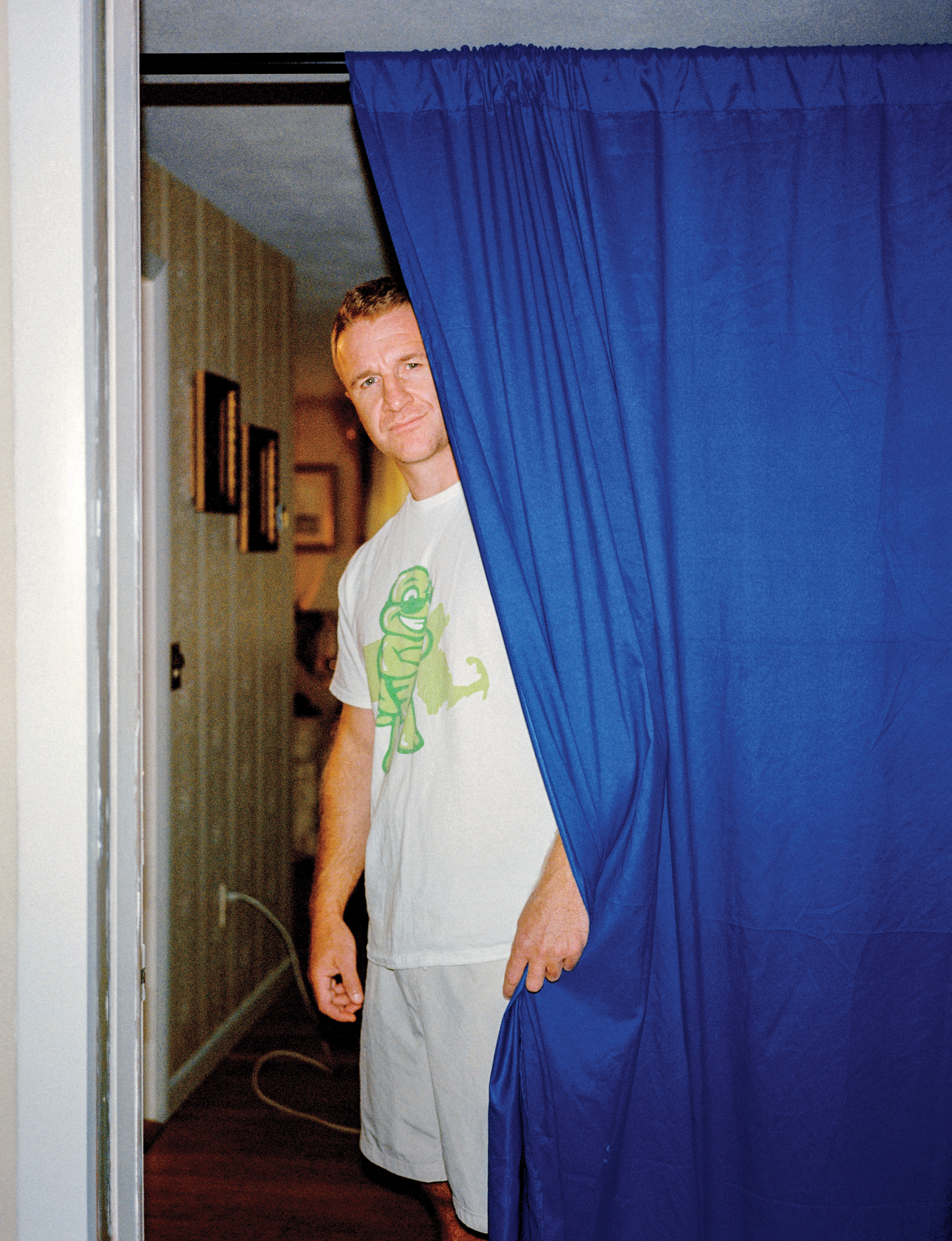 photo of man wearing white t-shirt with turtle logo and shorts looks out from behind a blue curtain inside a home