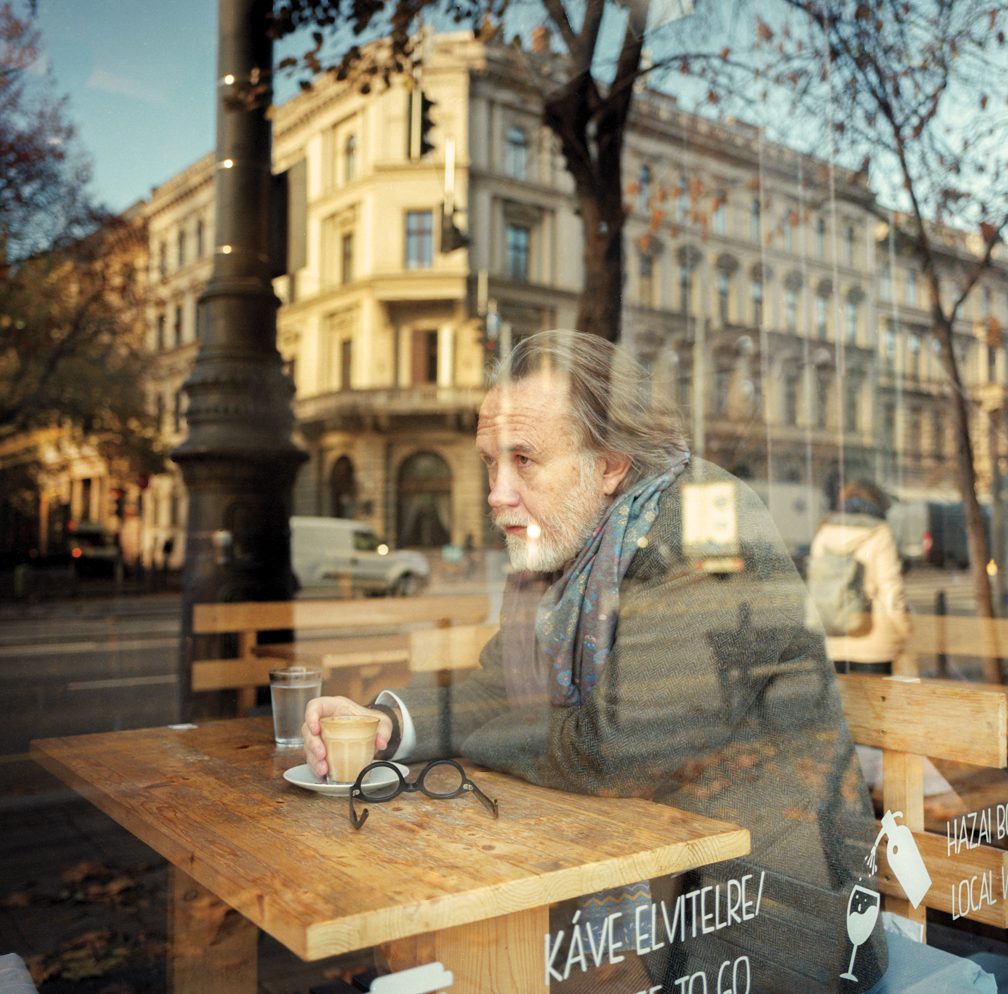 a portrait of Rod Dreher, wearing a gray coat and large scarf, sitting at a cafe table in Budapest