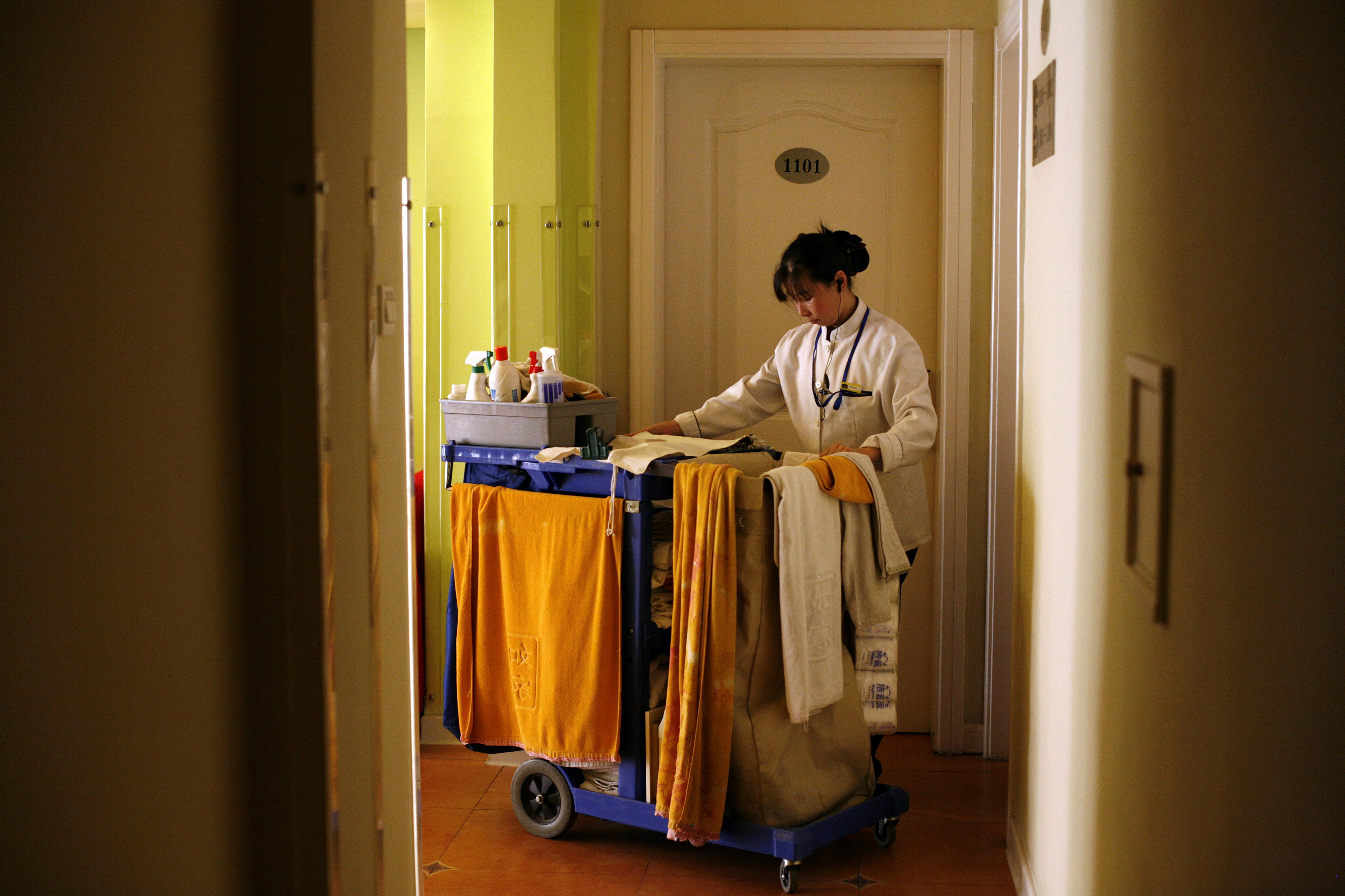 A hotel maid enters a room with a cart of cleaning supplies.