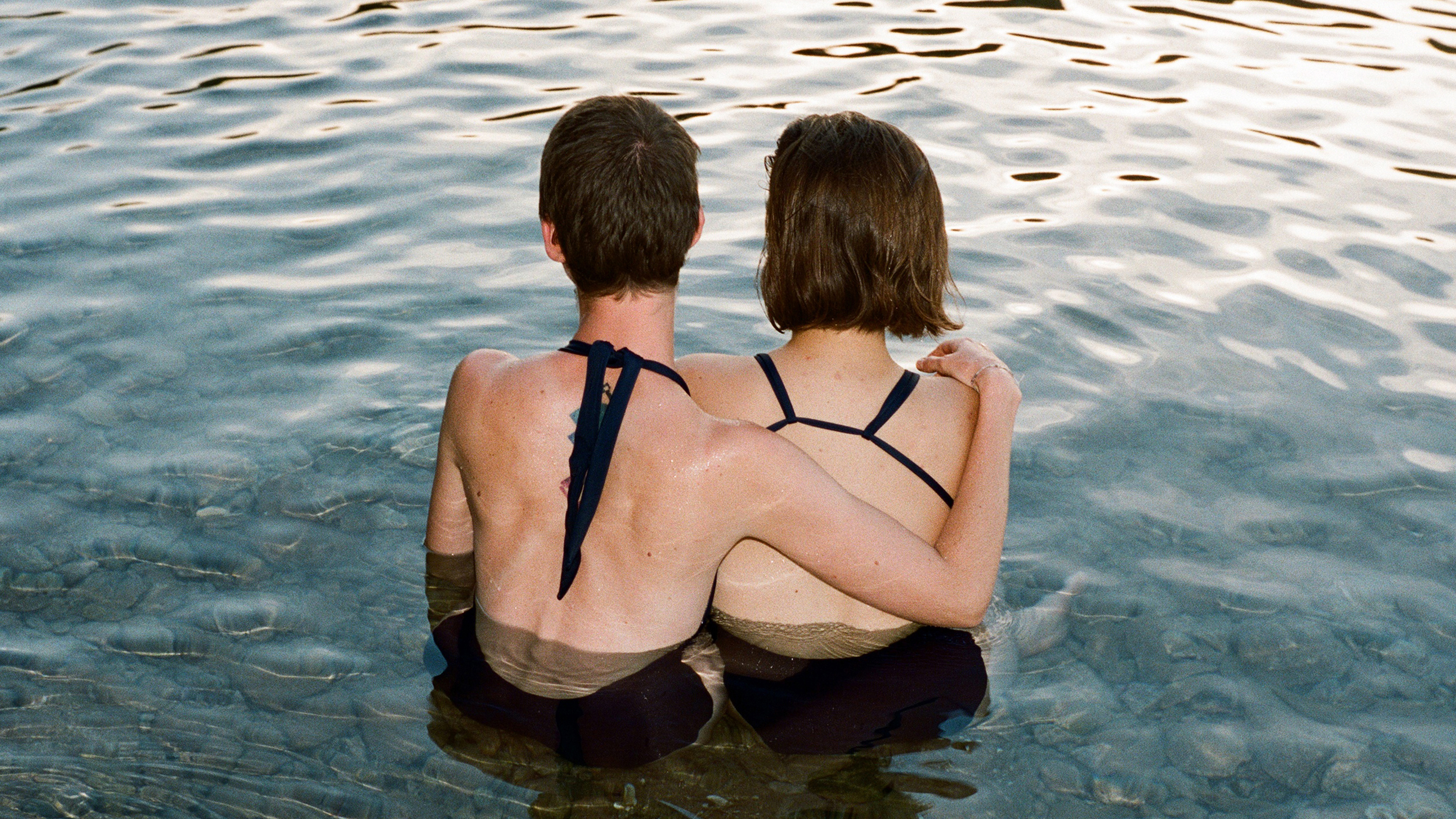 two people in bathing suits in lake, facing away from the camera, with an arm around each other