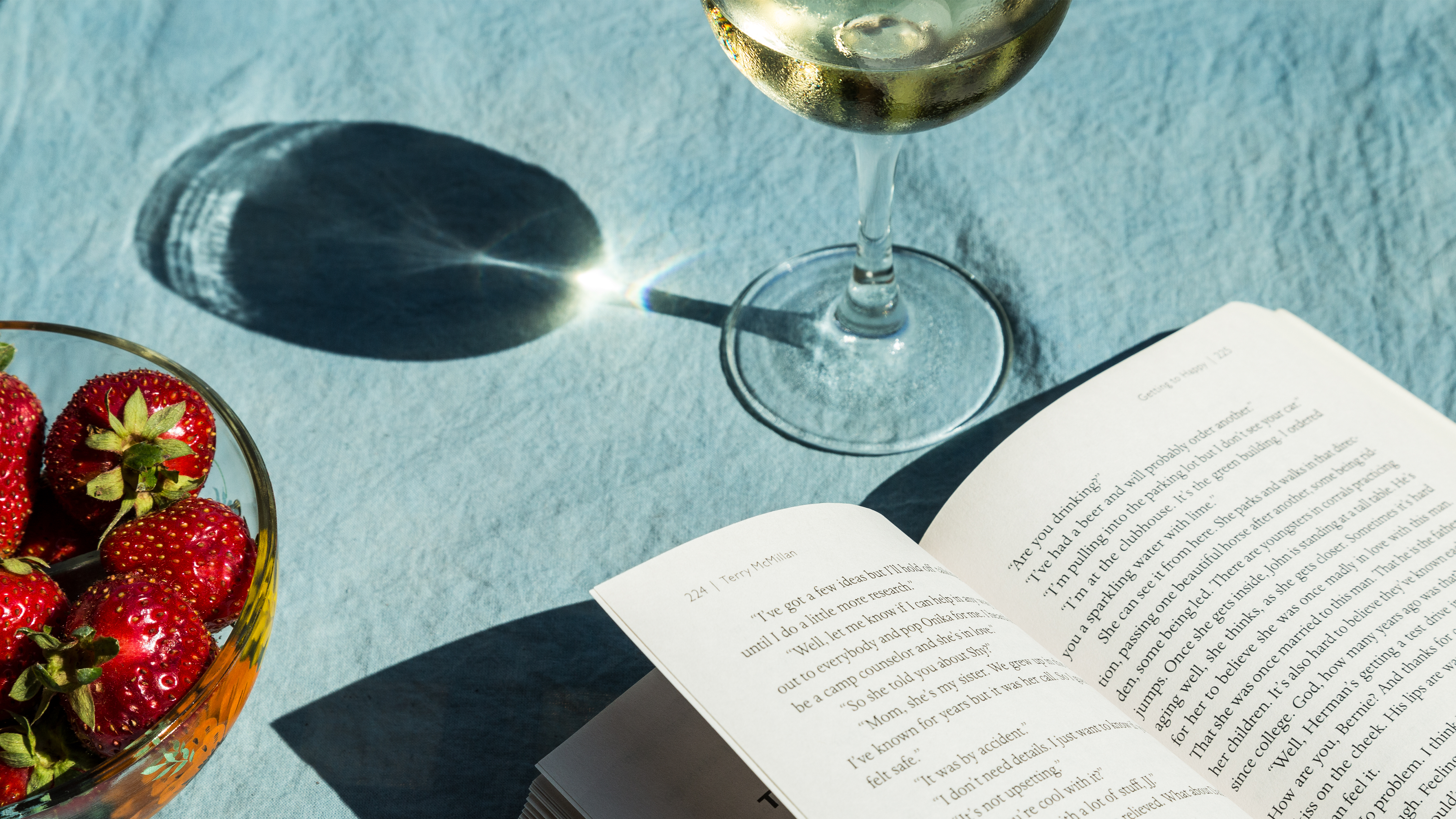 A bowl of strawberries, a glass of wine, and a book open on a tablecloth