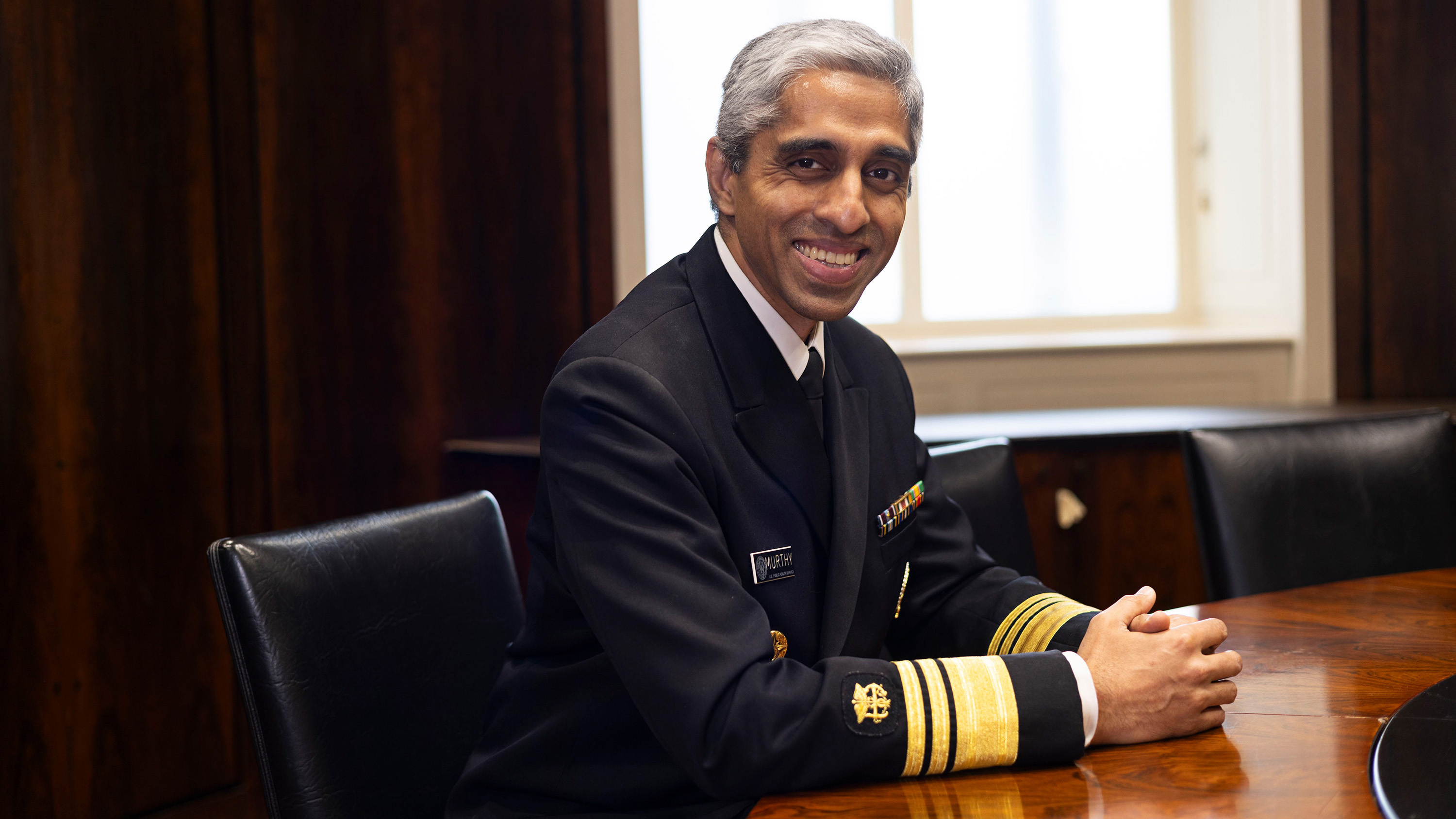 Surgeon General Vivek Murthy, in uniform, grins broadly and folds his hands on a shiny round table