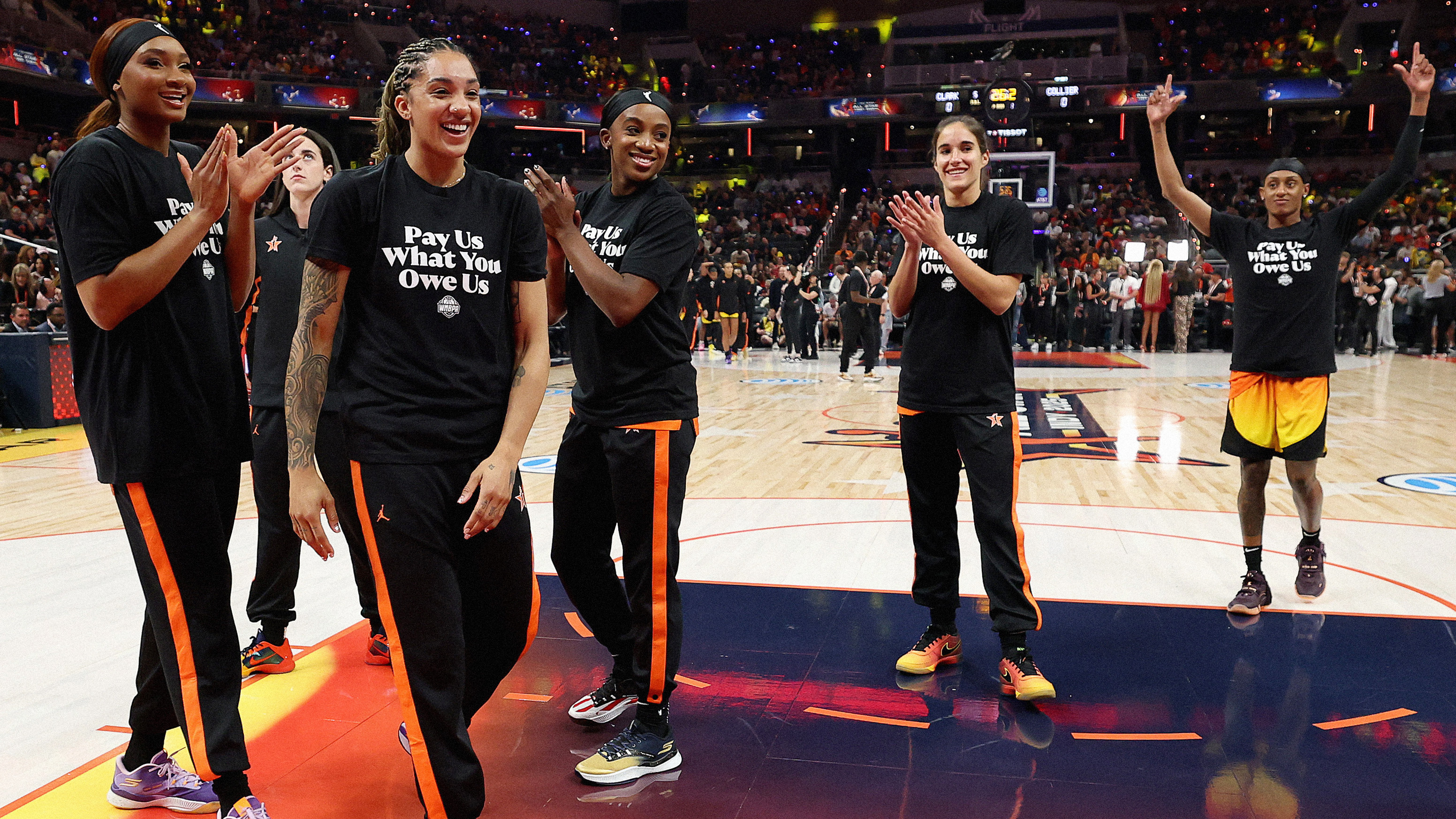 Photograph of WNBA athletes on a court wearing T-shirts that read “Pay Us What You Owe Us”