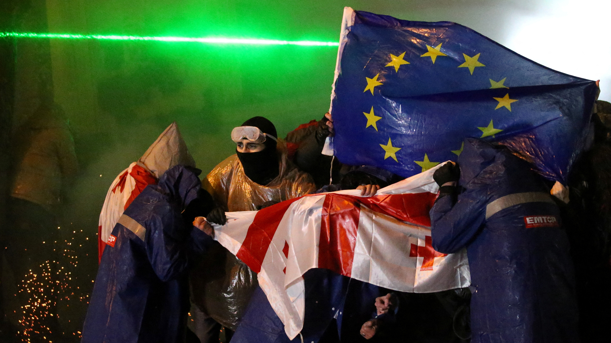 Photograph of protesters in masks surrounded by smoke, holding one flag of a red cross on white and another that is blue with a circle of yellow stars.