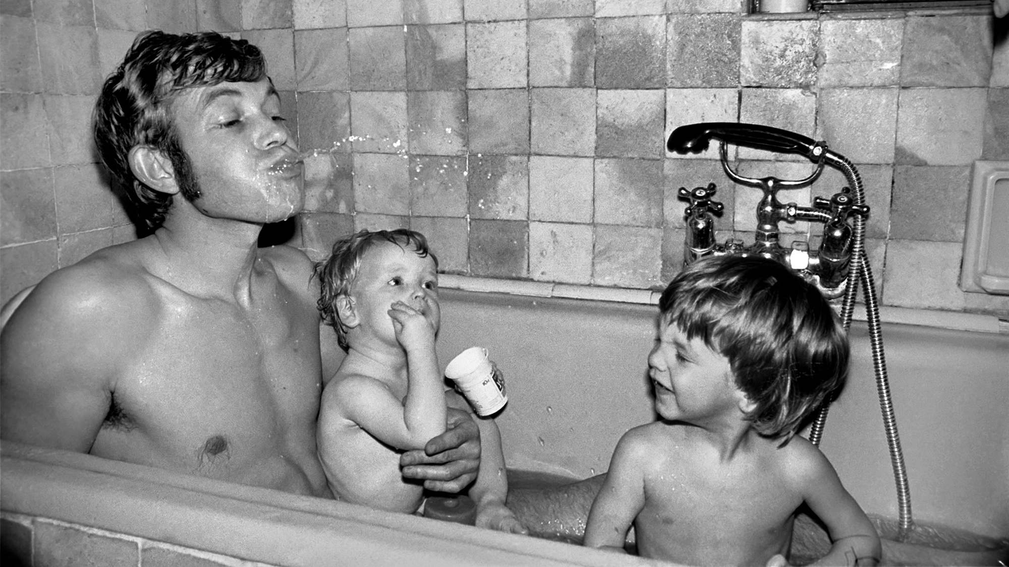 Black-and-white photograph of a dad and two sons in the bath, the dad spitting water out of his mouth