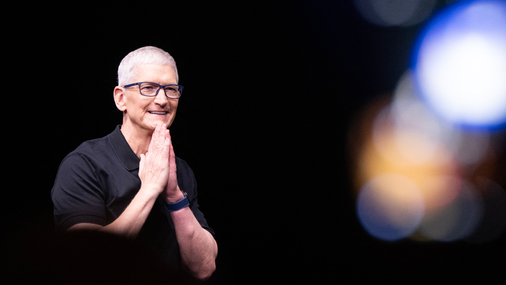 Tim Cook puts his hands together, touching his chin to both of them. Blurred lights in the right part of the background