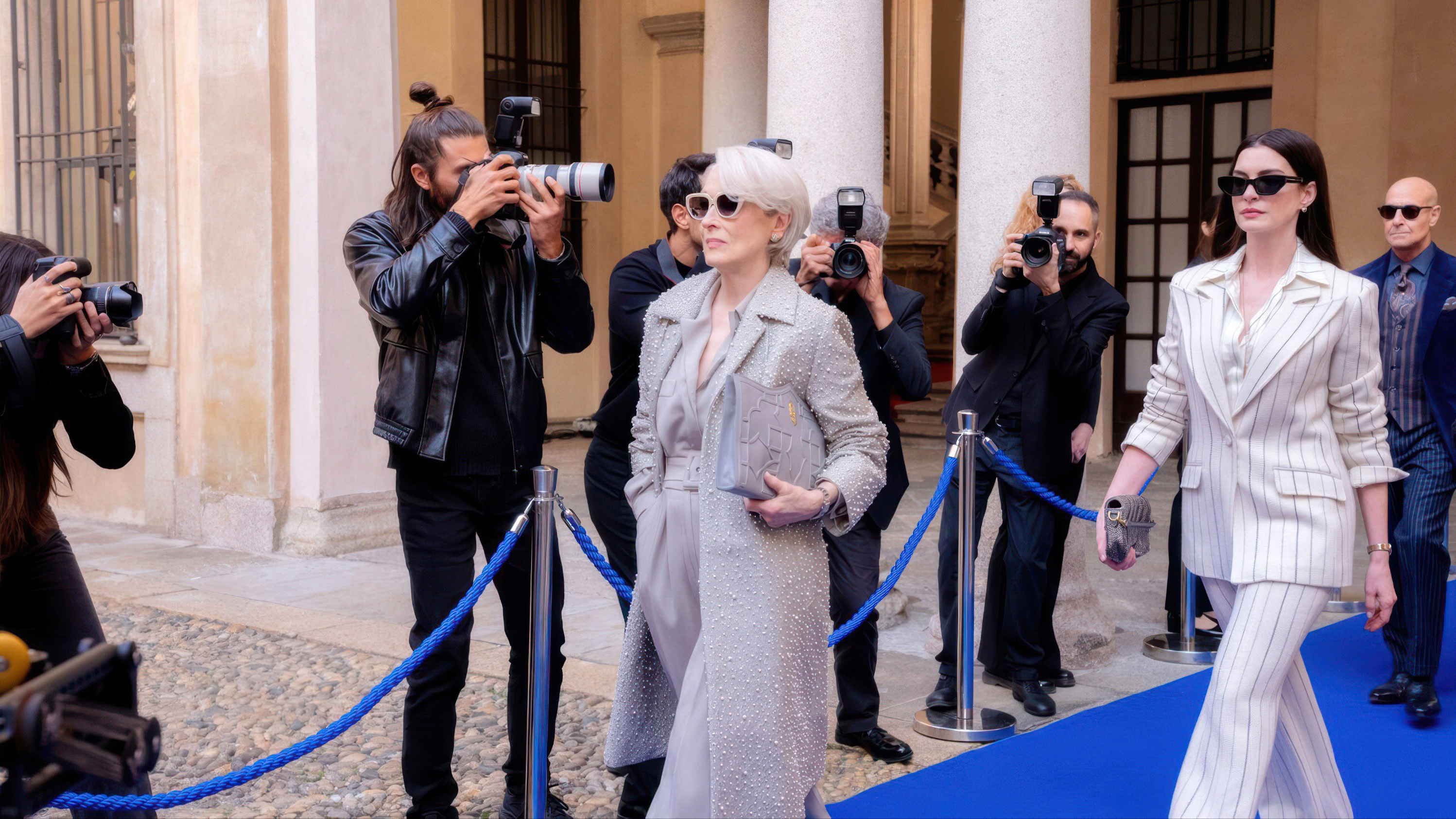 Meryl Streep and Anne Hathway, both dressed in silver, walk a blue carpet while being photographed in 'The Devil Wears Prada 2'