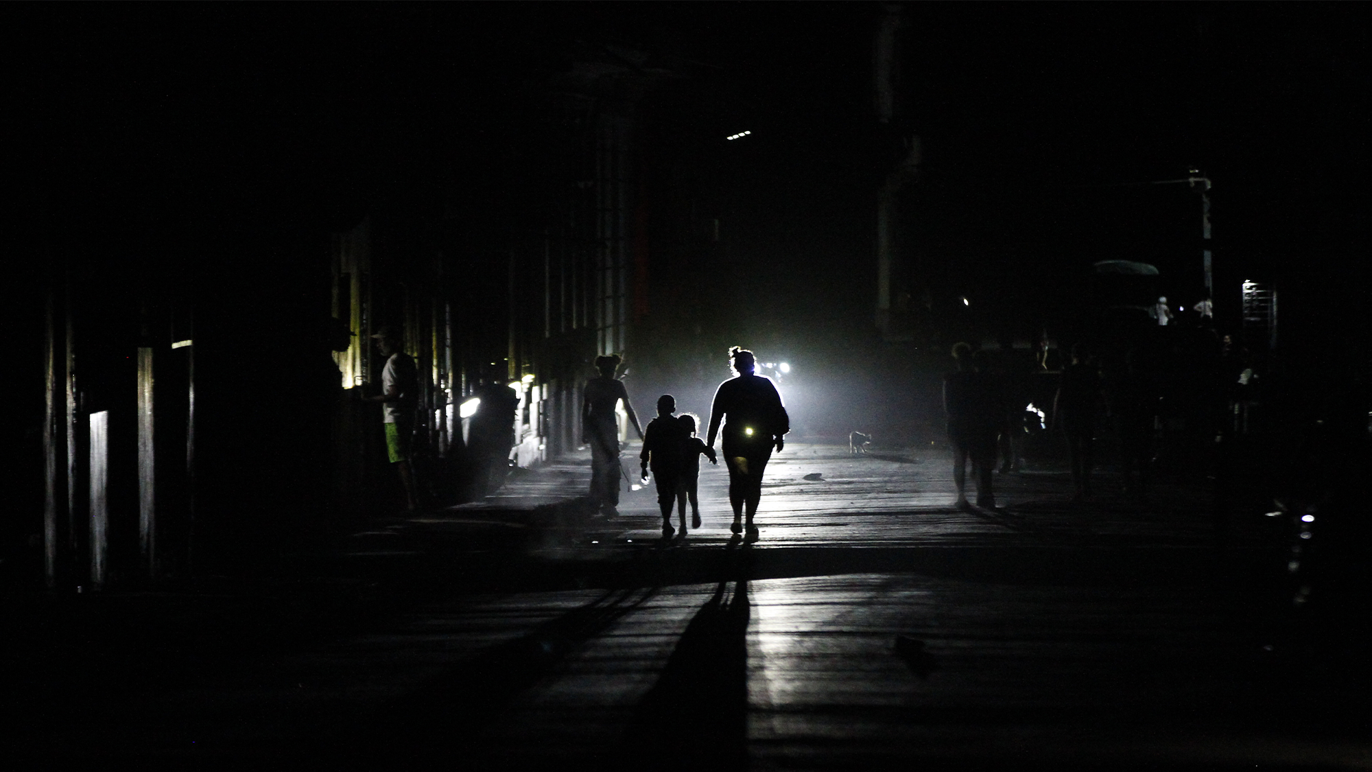 Color photo of a Cuban street in the dark, showing a the silhouettes of people lit up by headlights.