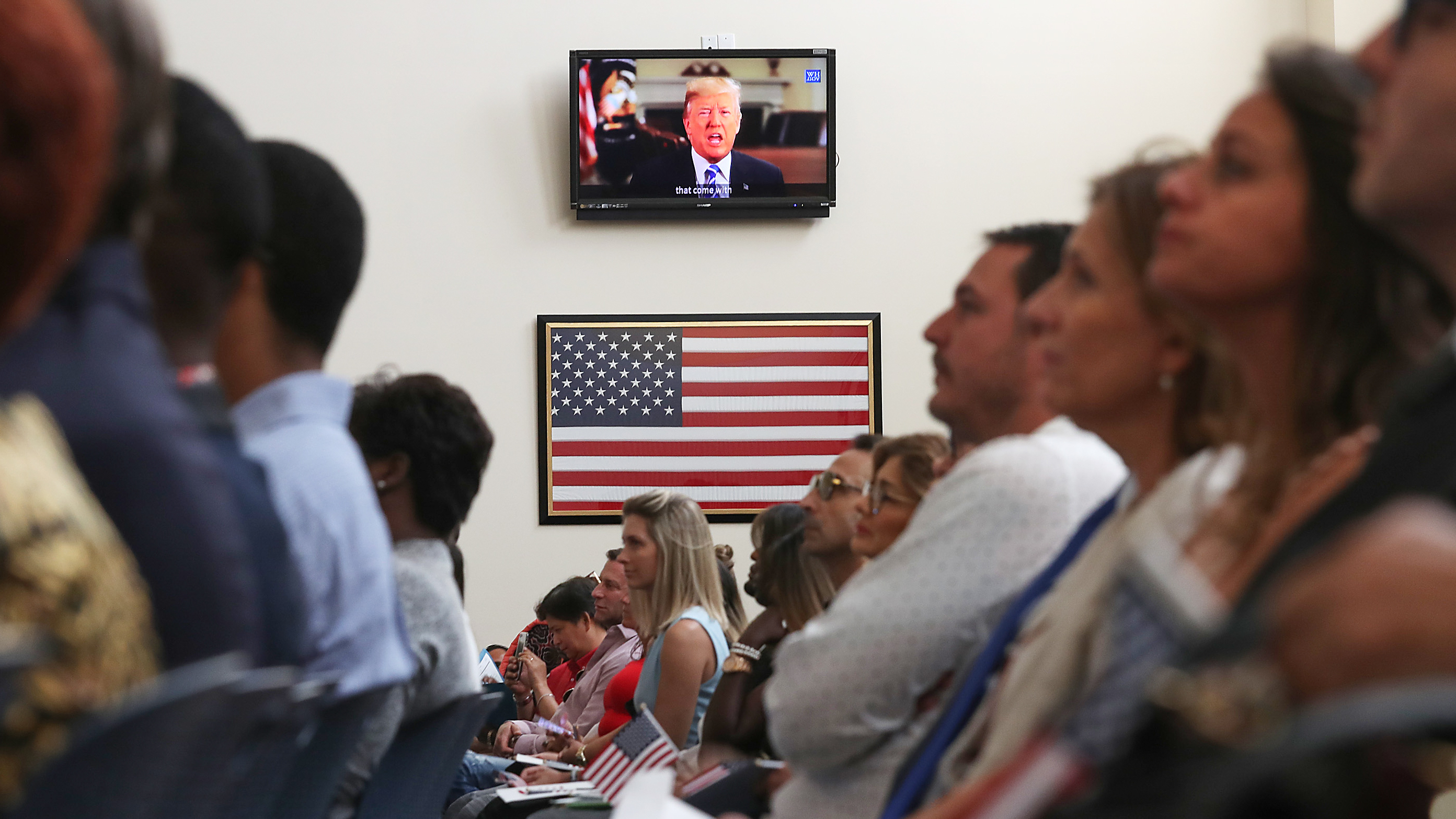 A crowd of people sitting look ahead while the camera focuses on an American flag and on a television screen with a video of Donald Trump speaking.