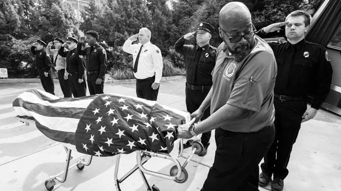 A man wheels out a guerney. A body on the guerney covered by a flag and first responders salute in the background. 