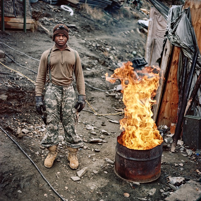 Soldier wearing hat, gloves, boots stands near half an oil drum with high flames burning