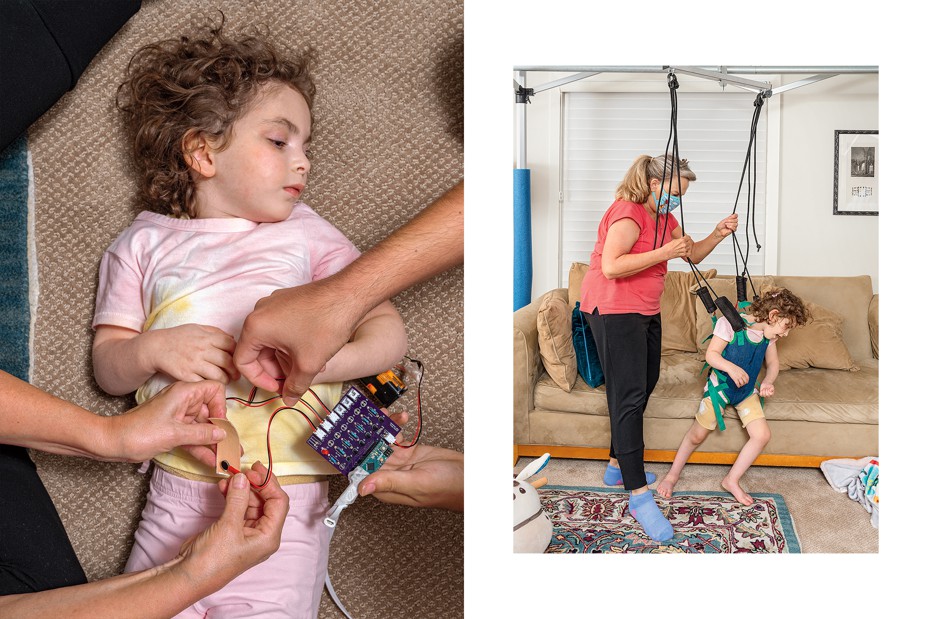 Livie lying down with head turned to side, viewed from above as one person holds a circuit-board with wires and battery pack and another holds an electrode. A woman with a ponytail wearing a face mask stands in stocking feet in front of a couch assisting Livie, wearing a harness suspended by cords from overhead steel supports, with walking. 