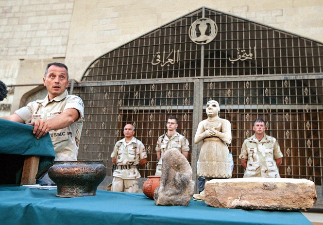 photo with Bogdanos at podium on left giving speech next to table with artifacts and statues in front of elaborate metal gates with 3 uniformed men in background