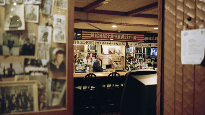 Photos are pinned to a bulletin board on the left side of this photo a looking through doors to a VFW bar. Wood panel doors are on the right side. A bartender is standing behind the bar in the middle of the photo.