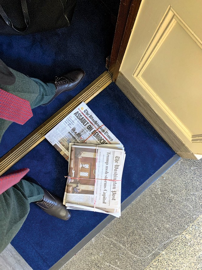 photo taken by Meijer looking down, showing the ends of his red tie, pants, and shoes standing on blue carpet next to several bundled newspapers