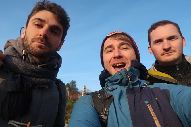 Three men dressed warmly smile for the camera, shoulder to shoulder in front of a bright-blue sky.