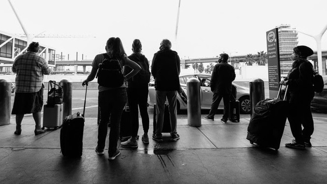 Black and white photo of people standing next to their suitcases, with their backs to the camera, outside an airport.