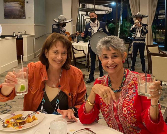 Two smiling women raise a glass to the camera while a mariachi band plays in the background