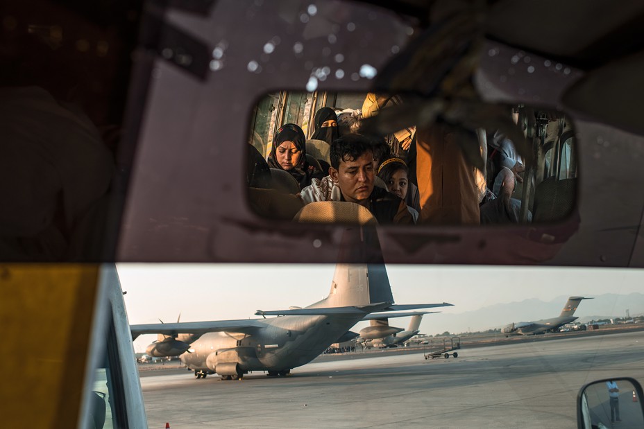 View out the front windshield of a bus facing military aircraft on an airfield, with the rear-view mirror showing the faces of people waiting inside it