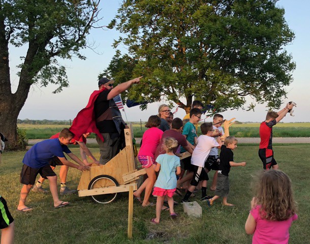 A man stands in a homemade chariot, wearing a cape and pointing forward. He's surrounded by children who are pushing and pulling the chariot.