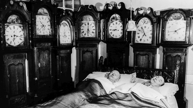 A man and woman sleep in a room full of antique clocks.