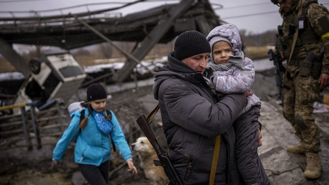 A man walks through a war zone in Ukraine while carrying a small girl. They are dressed in winter coats. Another girl, wearing a hat and a blue winter coat, walks behind them. A soldier wearing fatigues is visible on the right and an overturned car is shown in the background.